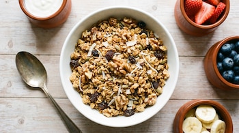 Conoce los 10 frutos secos con más proteína según la ciencia | 3 | Fotografía cenital de un bowl blanco con granola, nueces, semillas, pasas y coco, junto a una cuchara, yogur, frutillas, arándanos y banana en una mesa de madera.